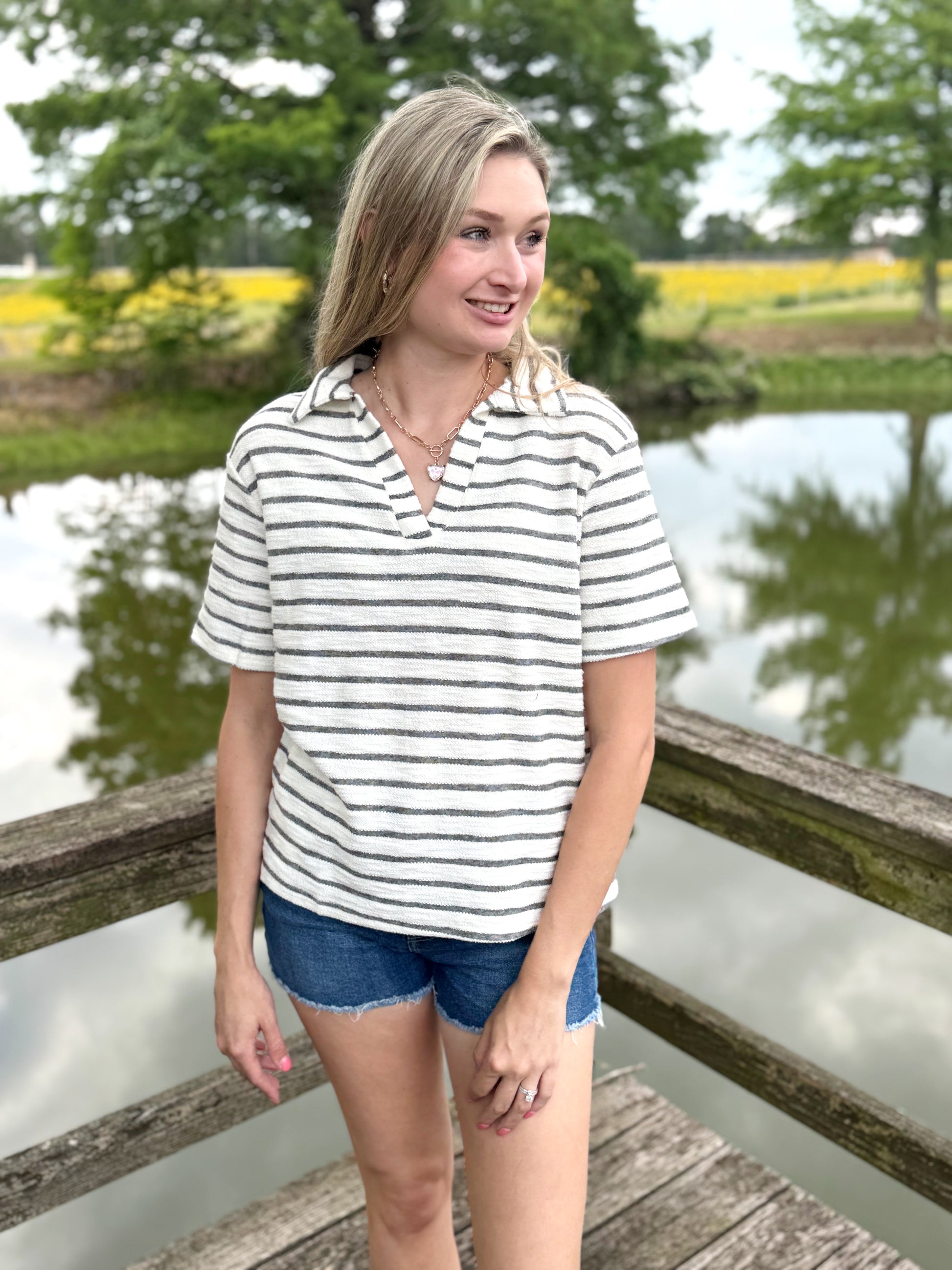 Woman in a striped shirt and denim shorts standing on a wooden dock by a pond with greenery in the background.